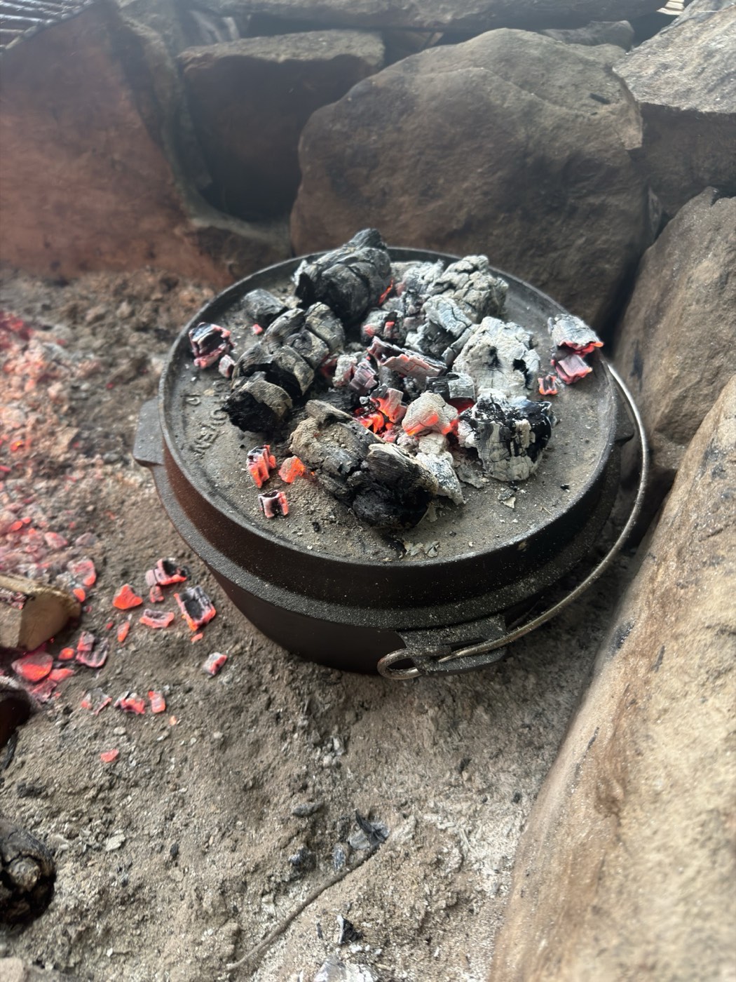 A cast-iron dutch oven covered in glowing coals, baking bread at a campsite fire pit