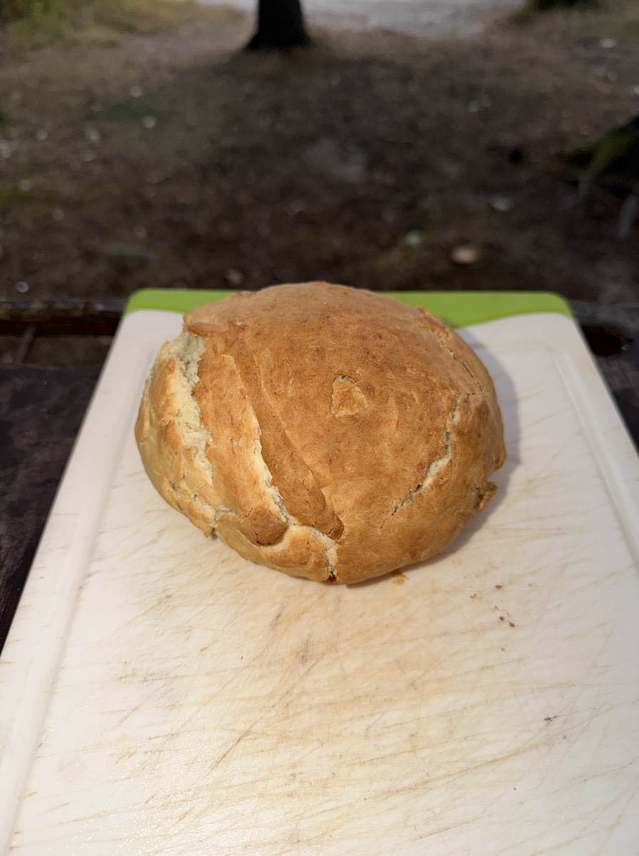 A golden loaf of bread baked in a dutch oven over a campfire