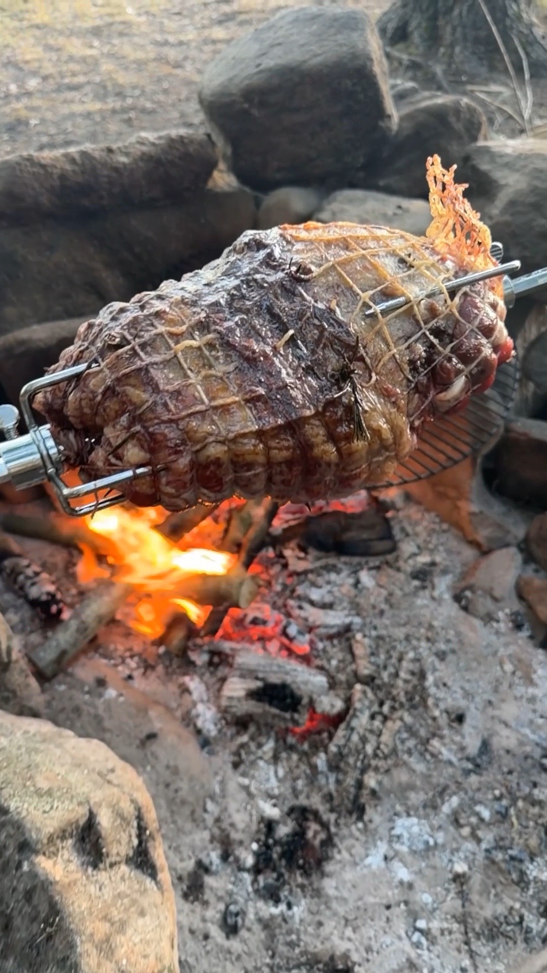 A leg of lamb turning on a spit over an open fire at a campsite