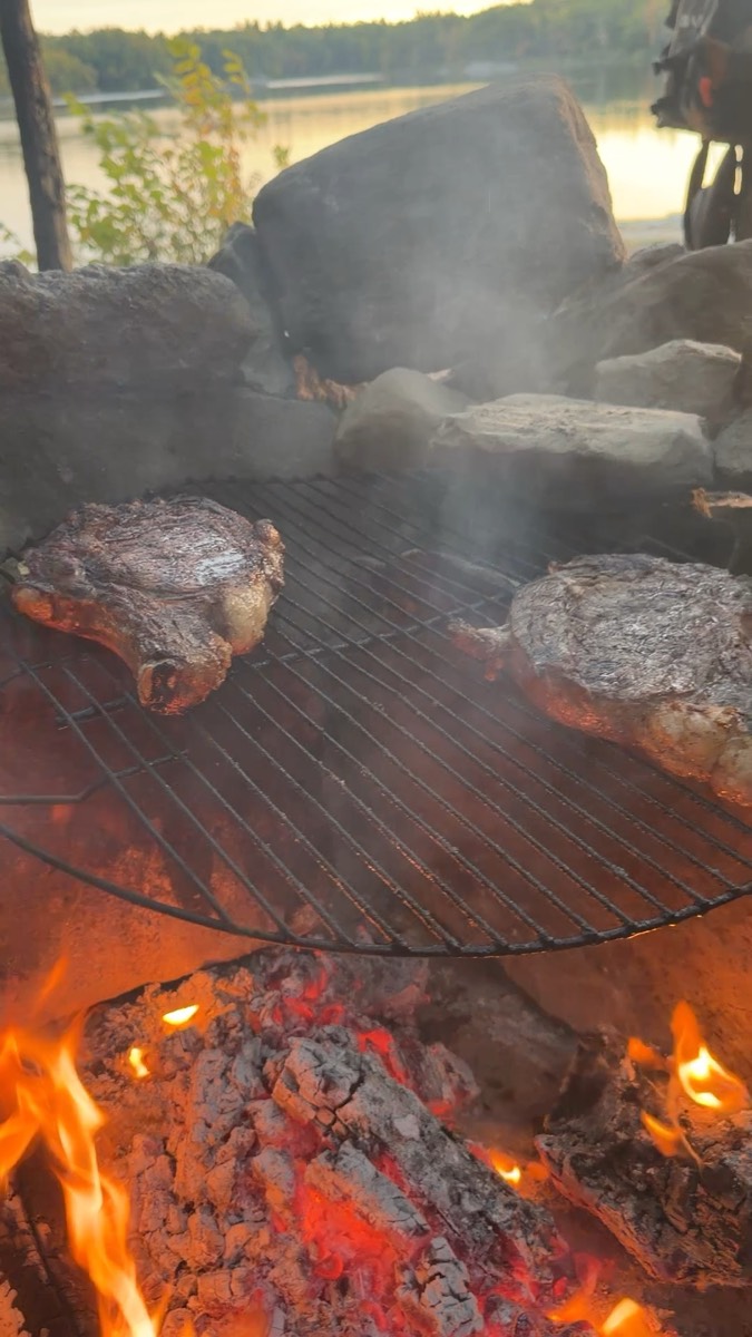 Two steaks grilling on a round grate over open flames at a lakeside campsite